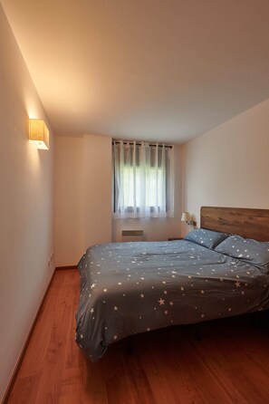 Ein Schlafzimmer mit einem Bett, einem Fenster mit Vorhängen, einer Wandlampe und einem Holzboden.