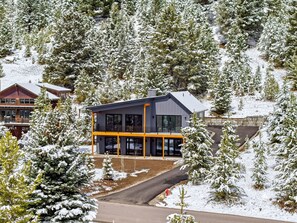 A modern two-story building with large windows and a balcony, surrounded by snow-covered trees. - Big Sky Sanctuary | Mtn Views & Deck & Hot Tub (Big Sky)