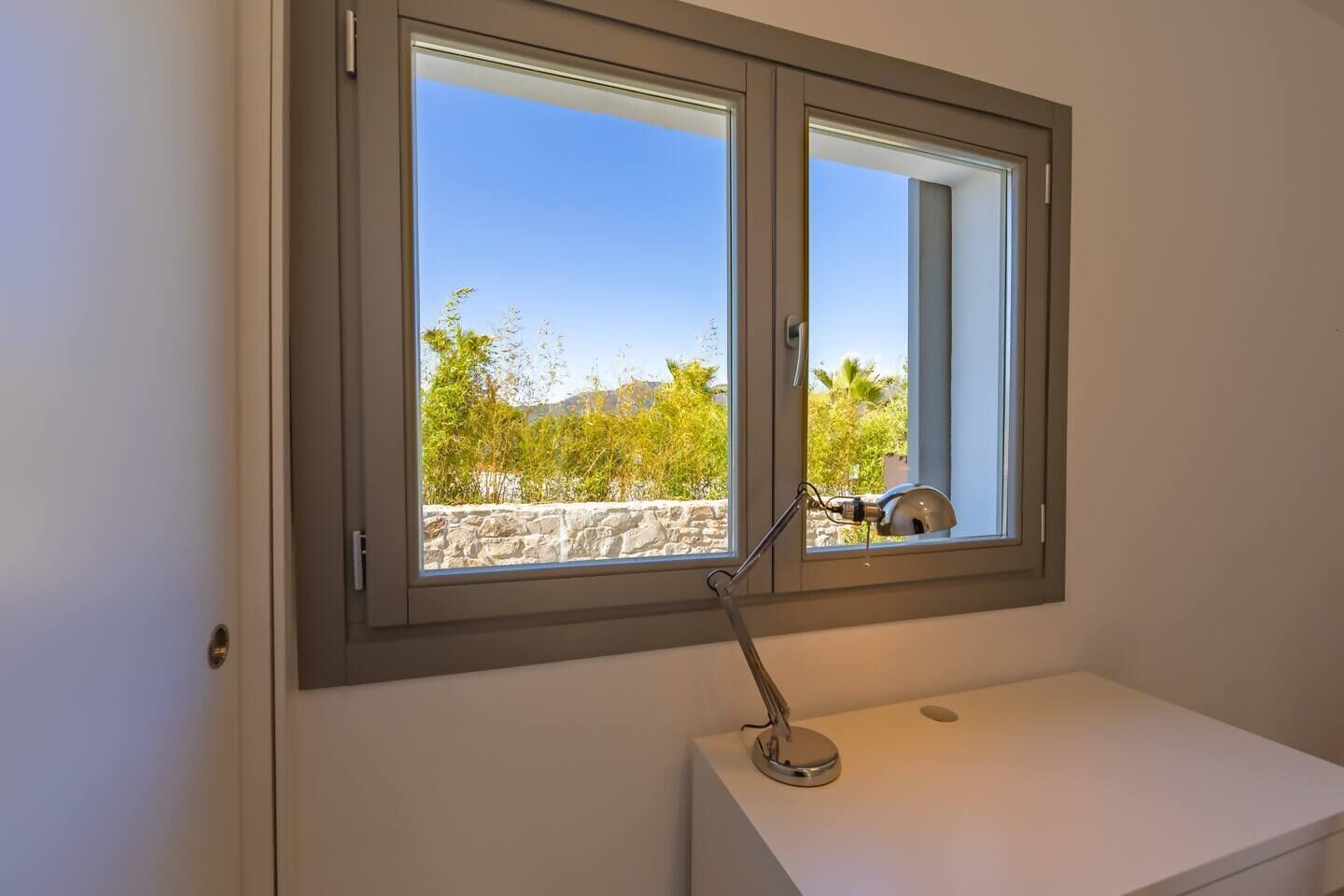 Una habitació amb escriptori blanc, una làmpada i una finestra amb vista a vegetació i una paret de pedra.