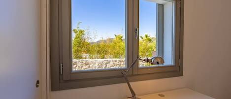 Una habitació amb escriptori blanc, una làmpada i una finestra amb vista a vegetació i una paret de pedra.