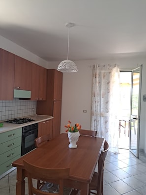 A kitchen with wooden cabinets, a gas stove, a white pendant light, and a dining table with chairs. - The Residences 1 (Marina di Modica)