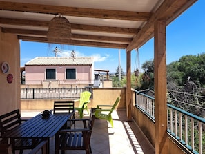 A covered outdoor area with a wooden table and chairs, a hanging light fixture, and a view of a building with a tiled roof. - The Residences 1 (Marina di Modica)