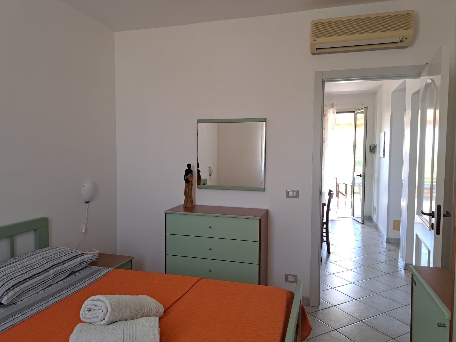 A bedroom with a bed, a dresser, a mirror, an air conditioner, and a hallway leading to another room.