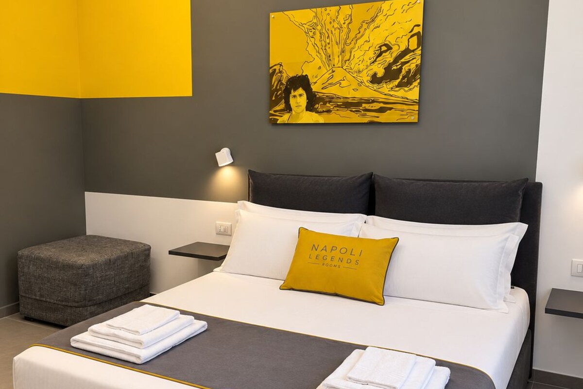 Een moderne slaapkamer met een bed, kussens, een nachtkastje, een lamp en een kunstwerk aan de muur.