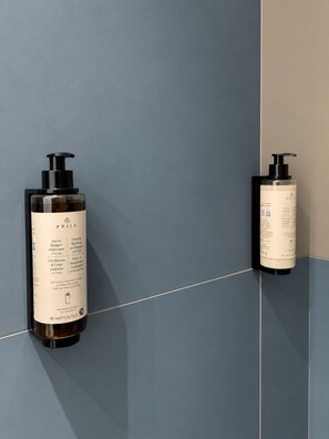 Twee flessen PRIJA shampoo en conditioner zijn aan een wand bevestigd, met een achtergrond van blauwe en beige tegels.