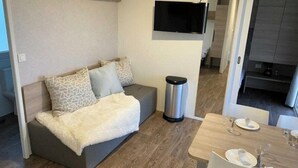 Ein kompakter Wohnbereich mit einer Couch, einer kleinen Mülltonne, einem Esstisch mit Besteck und einem Wand-Fernseher.