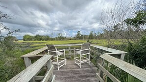 Une terrasse en bois avec deux chaises et une table, entourée de verdure et sous un ciel nuageux.