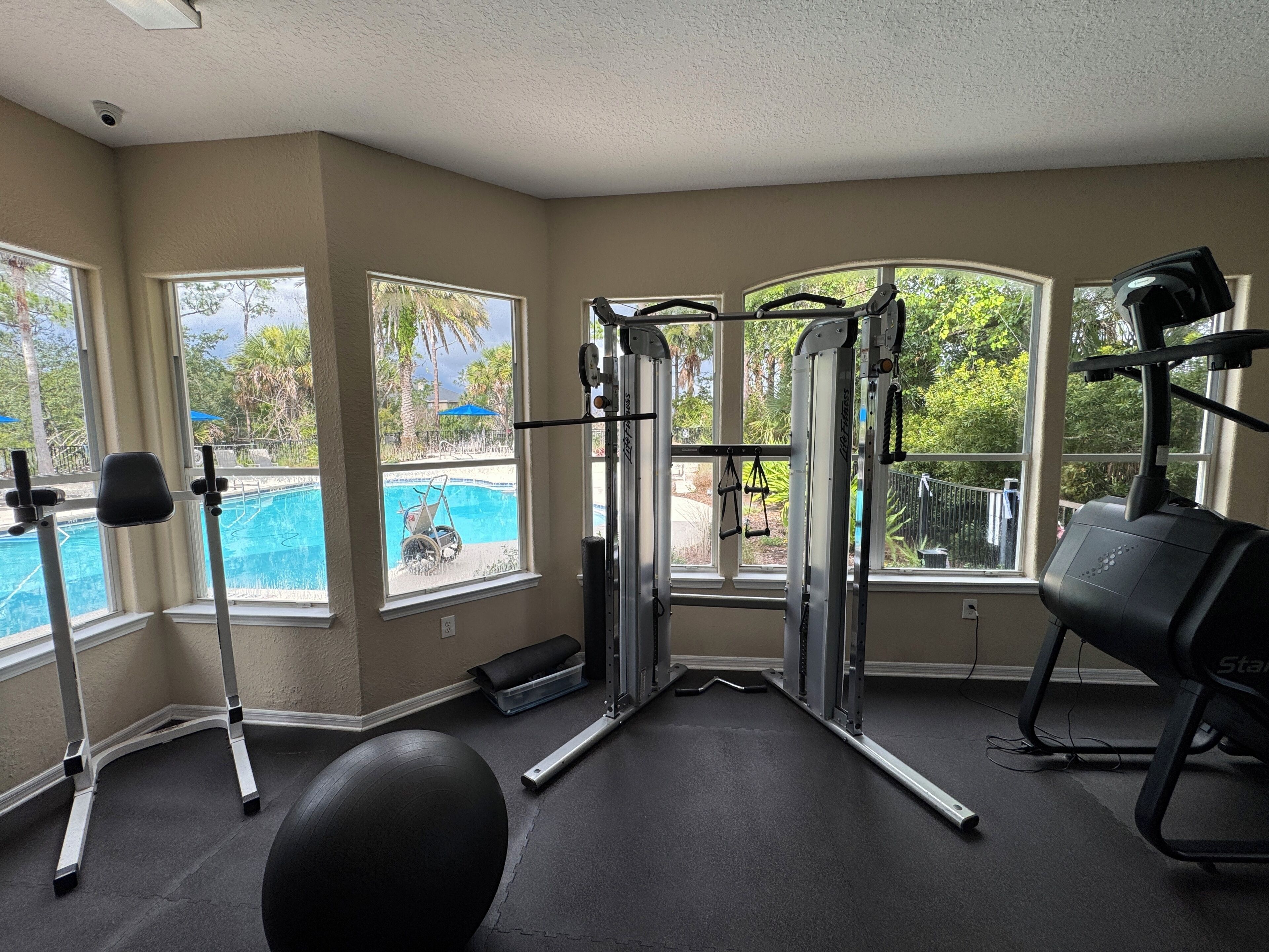 A home gym with exercise equipment, large windows, and a view of a swimming pool.