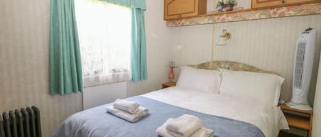 A hotel room with a bed, a radiator, a window with curtains, a bedside table, and a wall-mounted heater.