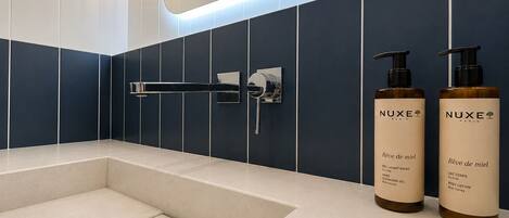 A modern bathroom with a bathtub, a hand shower, and two Nuxe Paris body lotion bottles.