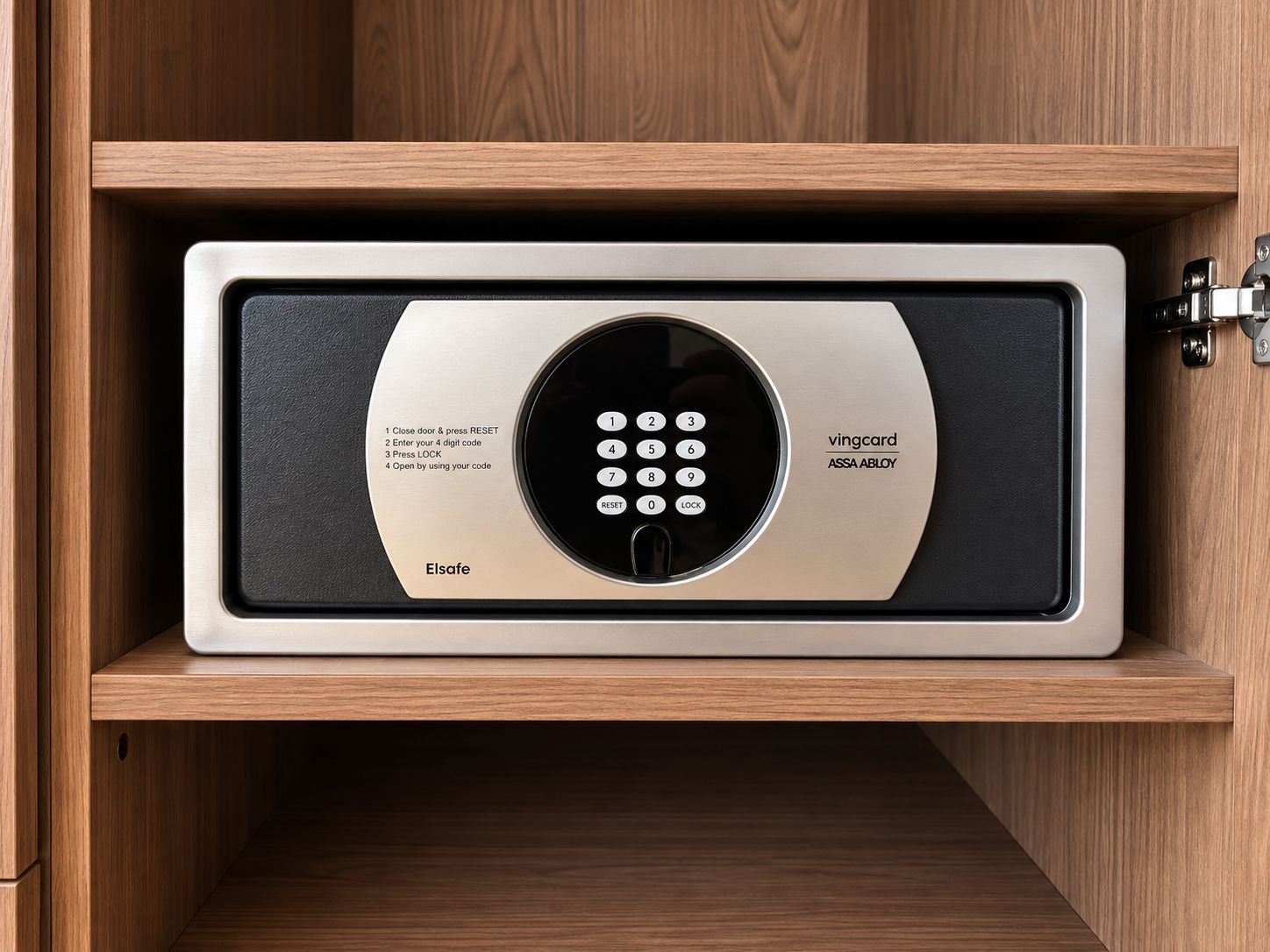 Una caja fuerte metálica segura con un teclado, etiquetada 'Vingcard ASSA ABLOY' y 'Elsafe', dentro de un mueble de madera.