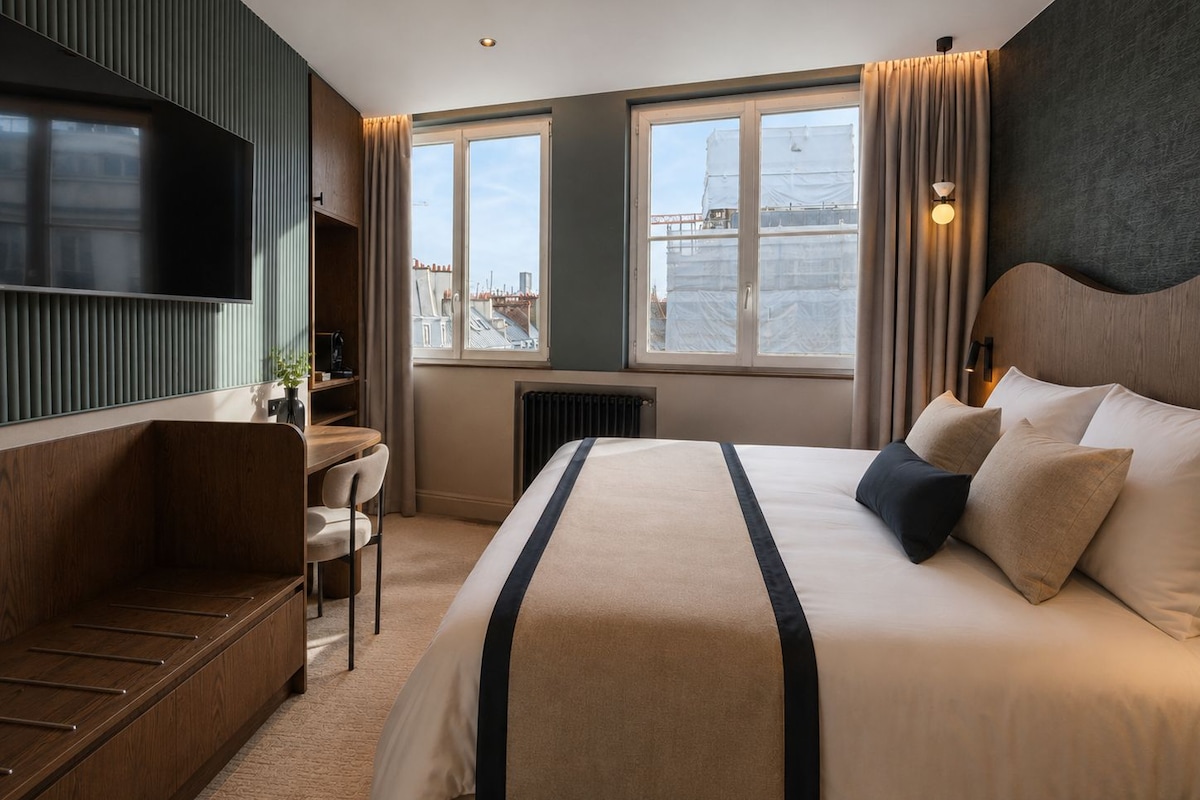 A hotel room with a bed, a desk, a chair, a TV, and a window with a view of buildings.
