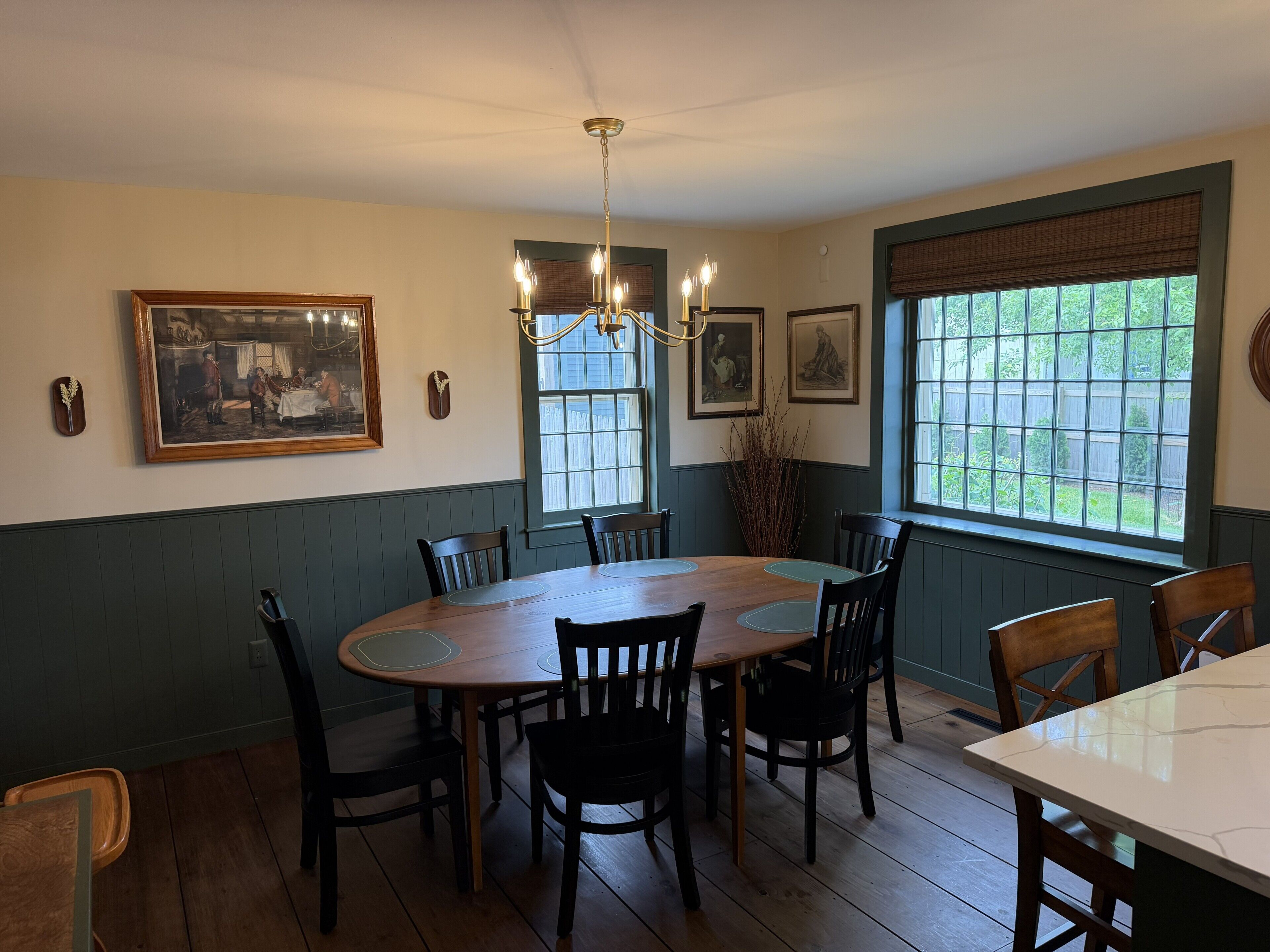 A dining room with a wooden table, chairs, a chandelier, framed artwork, and a window with a view.