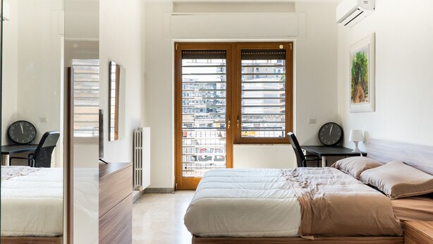 Ein Schlafzimmer mit Bett, Schminktisch, Schreibtisch, Stuhl und einem Fenster mit Blick auf Gebäude.