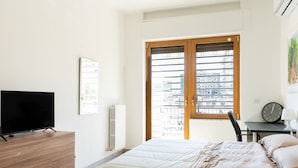 Ein Schlafzimmer mit einem Bett, einem Fernseher auf einer Kommode, einem Heizkörper, einem Fenster mit Jalousien und einem Bild an der Wand.