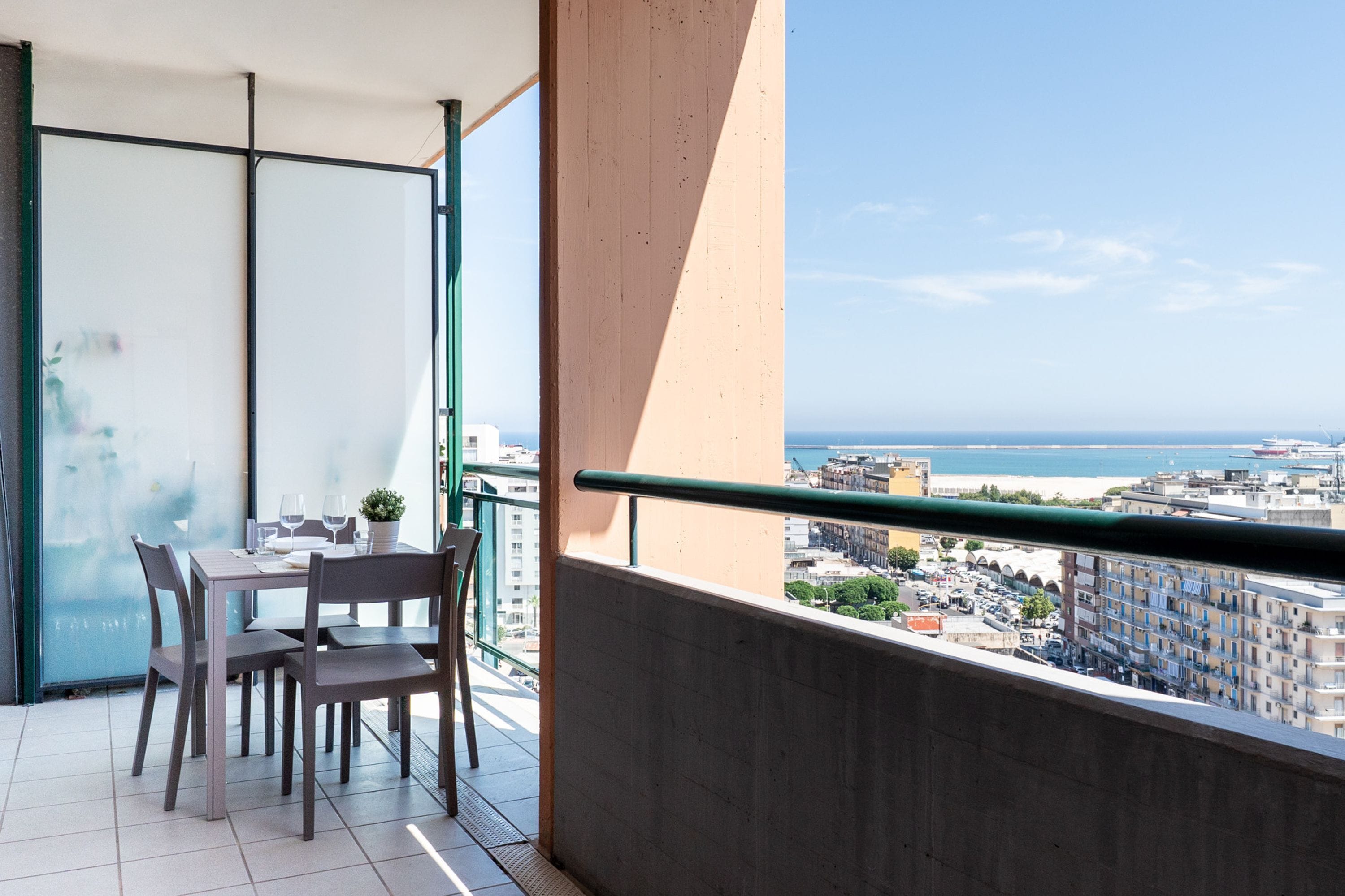 Een balkon met een eettafel en stoelen, een bloempot en uitzicht op zee en gebouwen.