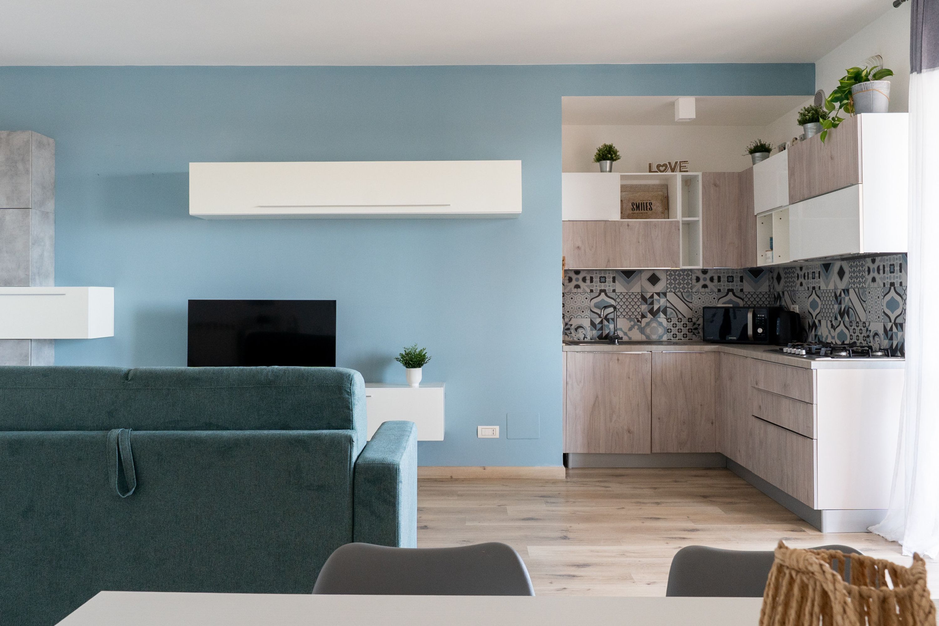 A modern living room with a sofa, a flat-screen TV, and a kitchen area with wooden cabinets and patterned backsplash.