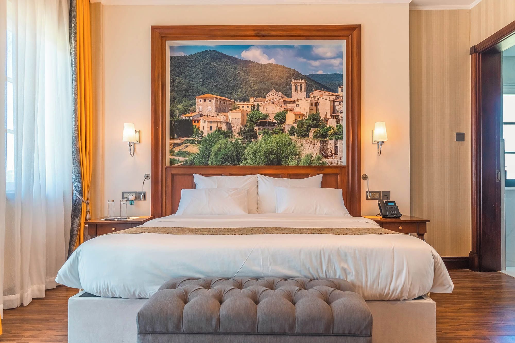A hotel room with a large bed, a headboard with a painting of a town, bedside lamps, a telephone, and a wooden floor.