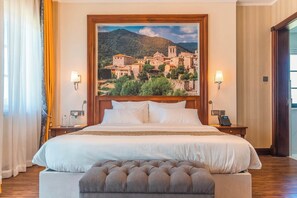 A hotel room with a large bed, a headboard with a painting of a town, bedside lamps, a telephone, and a wooden floor. - Avellino Lakeside Escape (Entebbe)