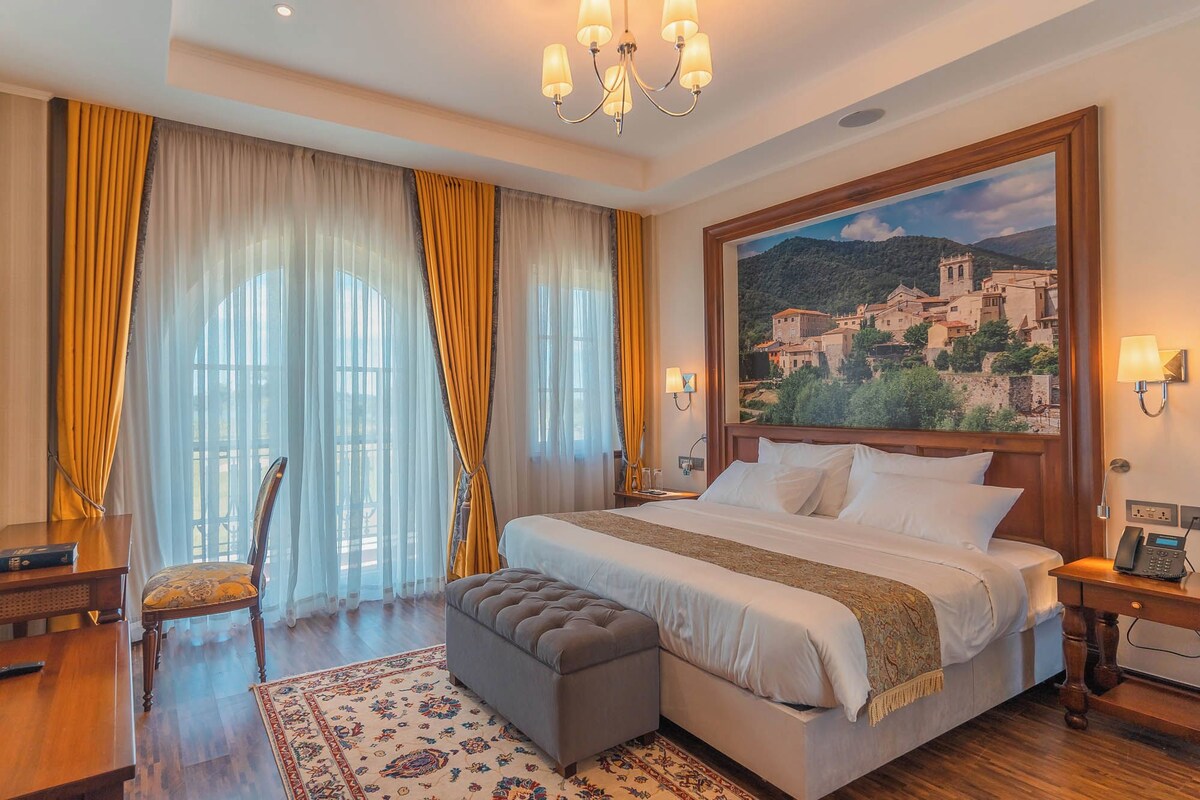 A hotel room with a large bed, a wooden nightstand, a desk with a chair, and a painting of a village on the wall.