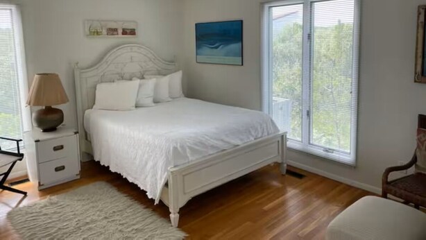 A bedroom with a white bed, a nightstand, a lamp, a chair, and a window with blinds.