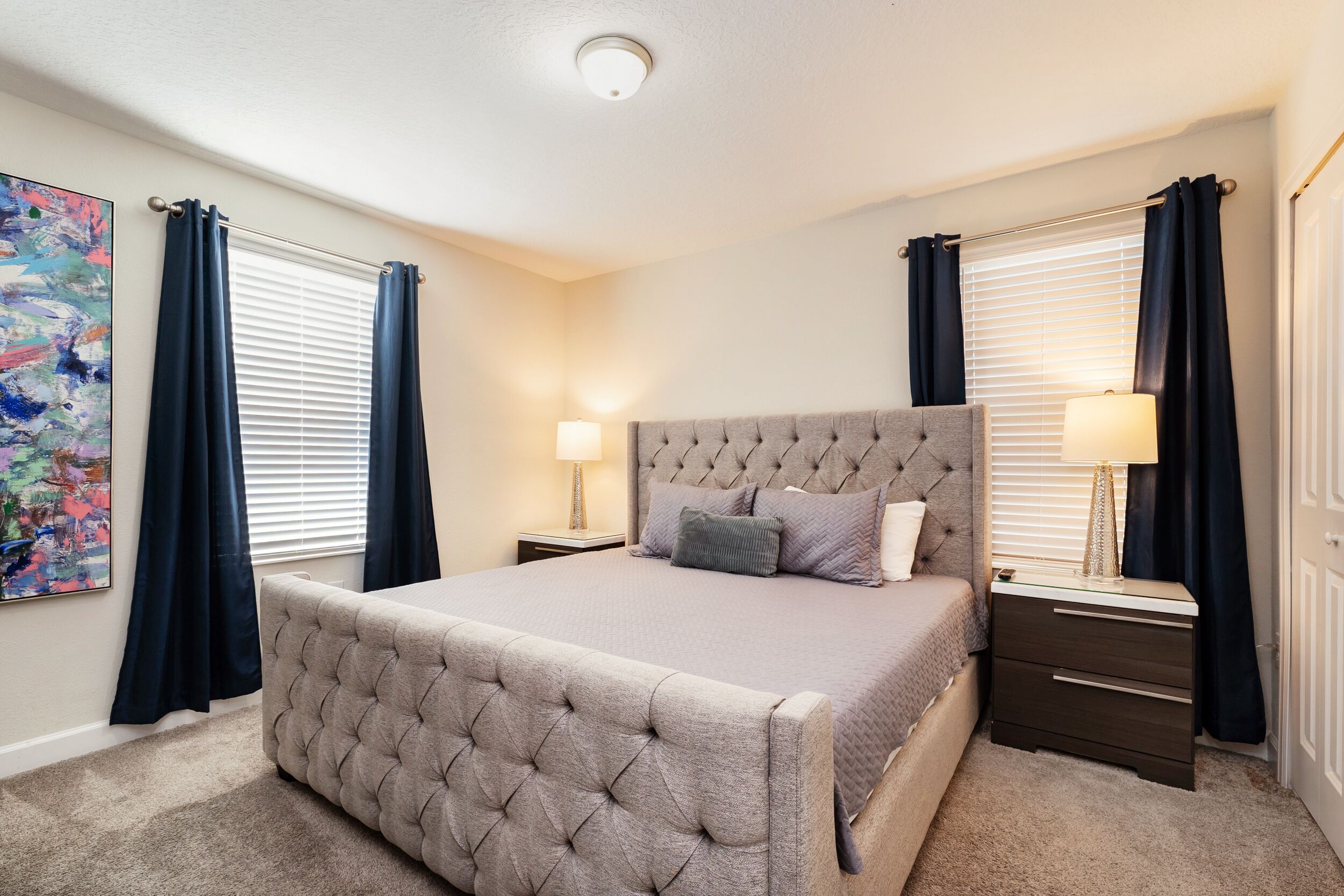 A bedroom with a tufted headboard bed, two bedside tables with lamps, and a large abstract painting on the wall.
