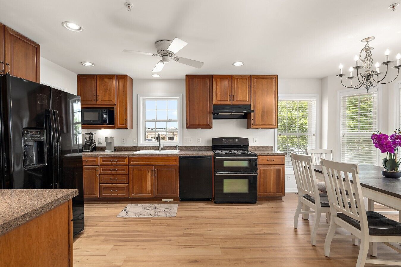Cocina moderna con gabinetes de madera, refrigerador negro, estufa y comedor con sillas blancas.