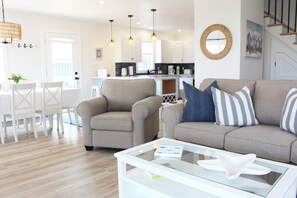 Ein modernes Wohnzimmer mit einer Couch, Sesseln, einem Glastisch und einem Essbereich mit weißem Tisch und Stühlen.