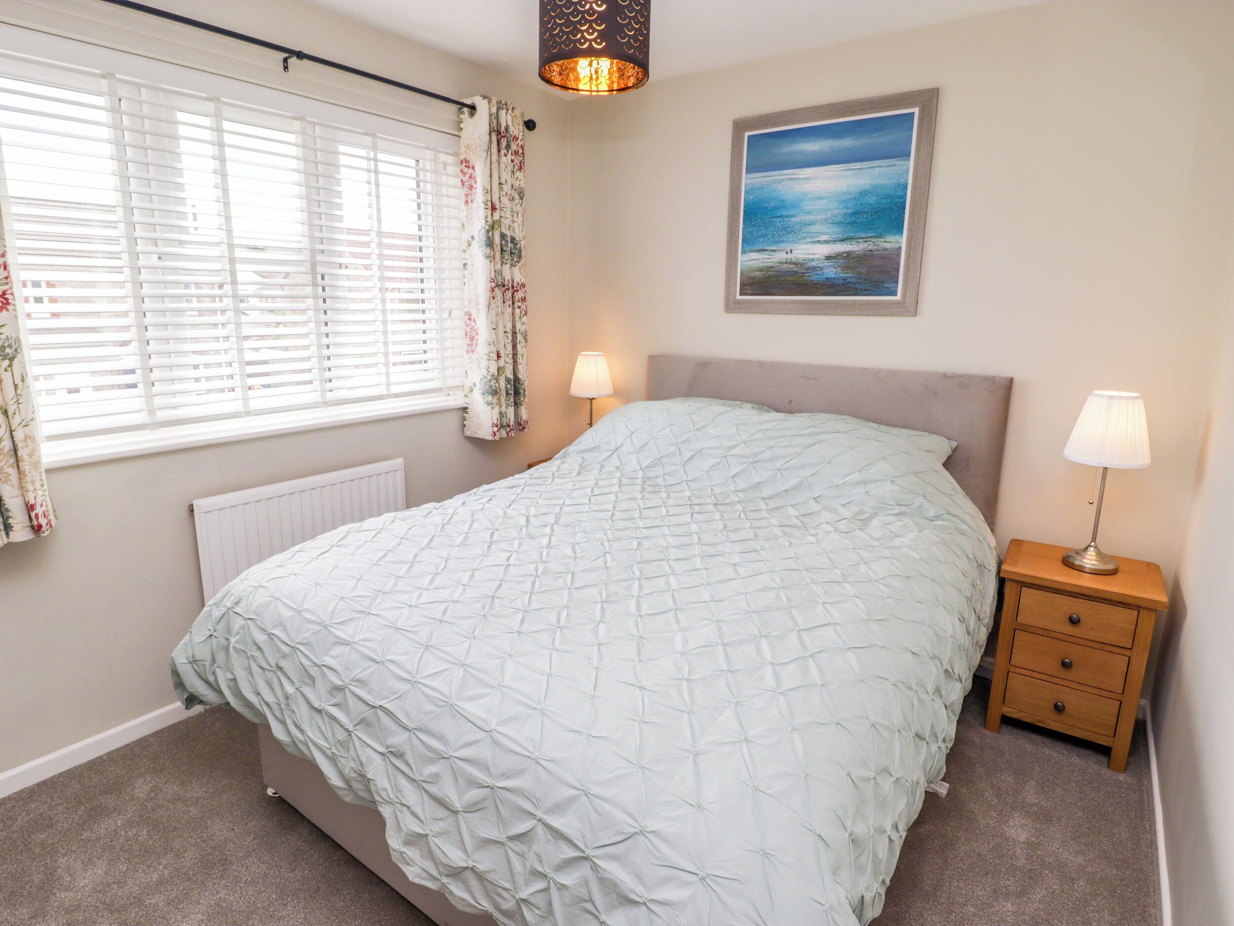 A bedroom with a bed, two bedside lamps, a radiator, a window with blinds, a floral curtain, and a framed picture on the wall.