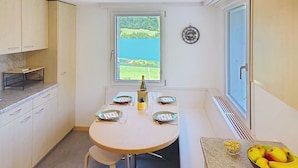 Et lille køkken med et spisebord dækket til fire personer, et vindue med udsigt til grøn natur, og et vægur.