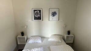 Una cama bien hecha con ropa blanca, dos mesitas de noche con lámparas y pequeñas plantas, y dos cuadros con motivos florales en la pared.