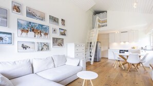 Ein zeitgemäßer Wohnraum mit weißem Sofa, Esstisch mit Stühlen, Küchenzeile und Schlafgalerie mit einer Leiter.