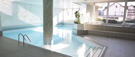 An indoor swimming pool with a clear view of the surrounding area, including a large column, tiled flooring, and a seating area with a view of buildings outside.