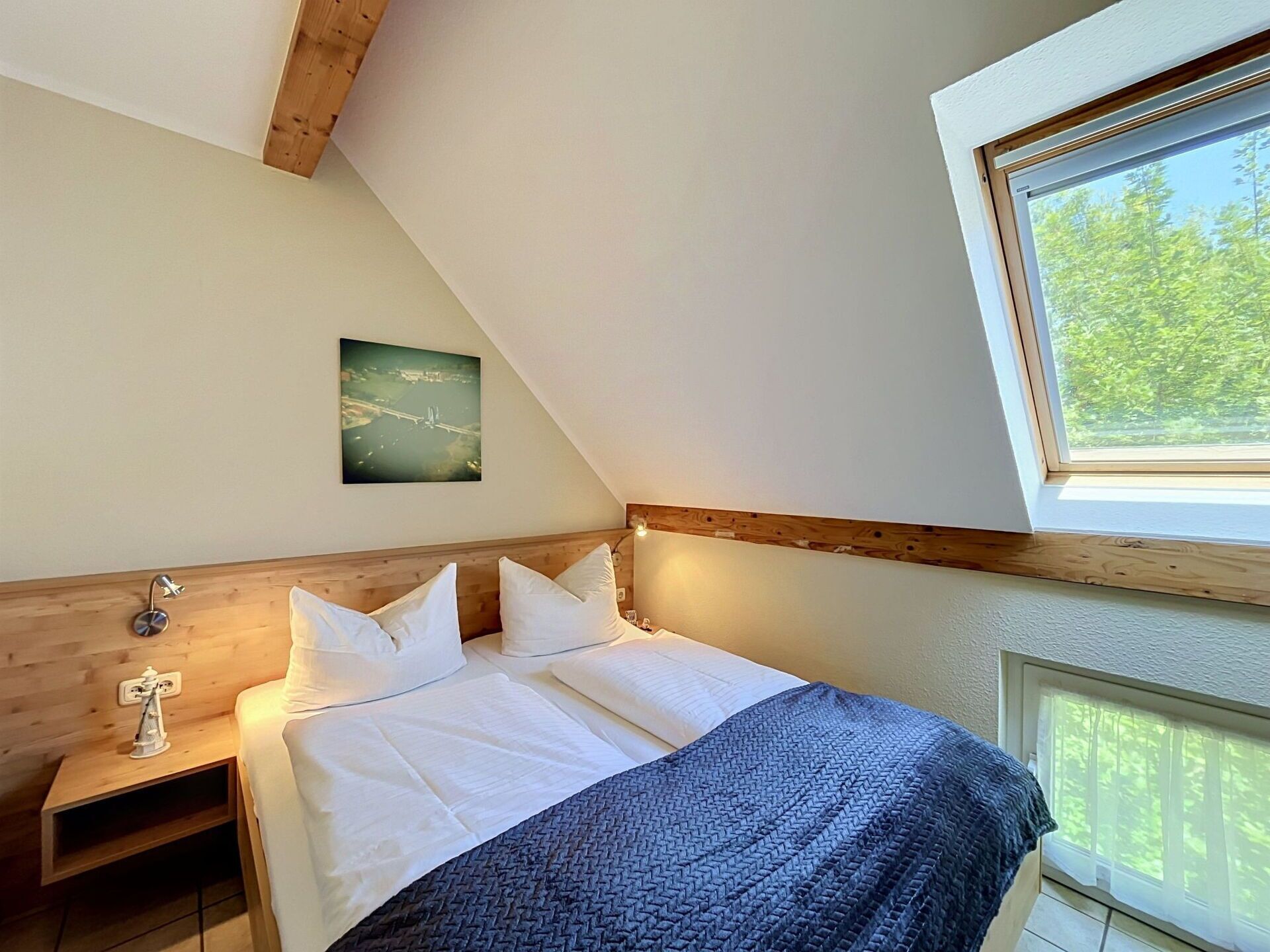 A bedroom with a wooden headboard, a bed with white bedding, a nightstand with a lamp, a framed picture on the wall, and a window with a view of trees.