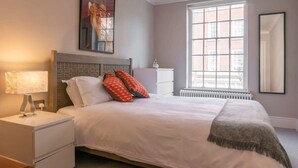 A bedroom with a bed, nightstand, lamp, window, radiator, and a painting. - A Regal Abode - Marylebone - by Frankie Says (London)