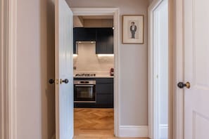 A hallway leading to a kitchen with dark cabinets, a stove, and an oven. - A Regal Abode - Marylebone - by Frankie Says (London)