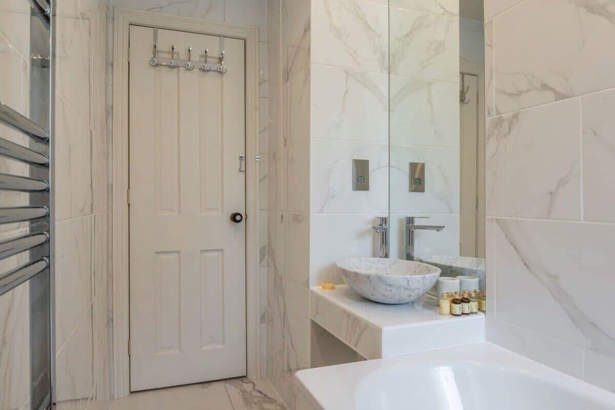 A bathroom with a white door, a marble-tiled wall, a large mirror, a sink with a marble basin, and a bathtub.