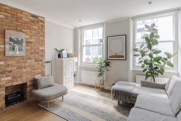A living room with a brick wall, a fireplace, a sofa, a chair, and two windows with plants. - Charming 2-bedroom Apartment in Fitzrovia (London)