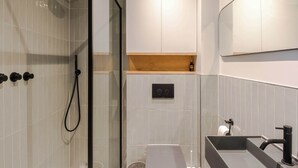 A modern bathroom with a glass shower enclosure, a built-in sink with a black faucet, and a wooden shelf above the sink. - Charming 2-bedroom Apartment in Fitzrovia (London)