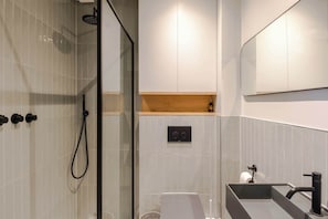 A modern bathroom with a glass shower enclosure, a built-in sink with a black faucet, and a wooden shelf above the sink. - Charming 2-bedroom Apartment in Fitzrovia (London)
