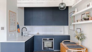 A modern kitchen with dark blue cabinets, a white countertop, a built-in oven, and a wooden dining table with two grey placemats. - Charming 2-bedroom Apartment in Fitzrovia (London)