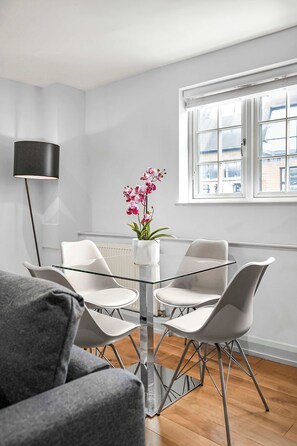 A modern dining area with a glass table, white chairs, and a vase of pink orchids. - Comfy 1-bed in Soho, Steps From Carnaby Street (London)