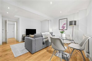 A modern living room with a grey sofa, a glass dining table with chairs, a floor lamp, and a flat-screen TV. - Comfy 1-bed in Soho, Steps From Carnaby Street (London)