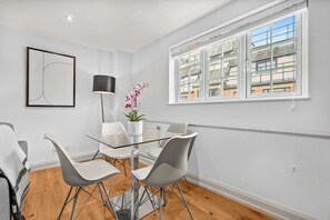 Een moderne eetruimte met een glazen tafel en witte stoelen, een potorkidee en een lijst met abstracte kunst aan de muur.