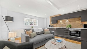 A modern living room with a grey sofa, a round coffee table, and a kitchen with dark cabinets and a brick backsplash. - Comfy 1-bed in Soho, Steps From Carnaby Street (London)