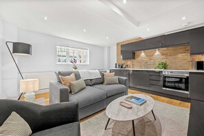 Een moderne woonkamer met een grijze bank, een ronde salontafel en een keuken met donkere kastjes en een tegelwand van baksteen.