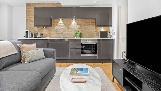 A modern kitchen with dark cabinets, a built-in oven, and a large TV. - Comfy 1-bed in Soho, Steps From Carnaby Street (London)