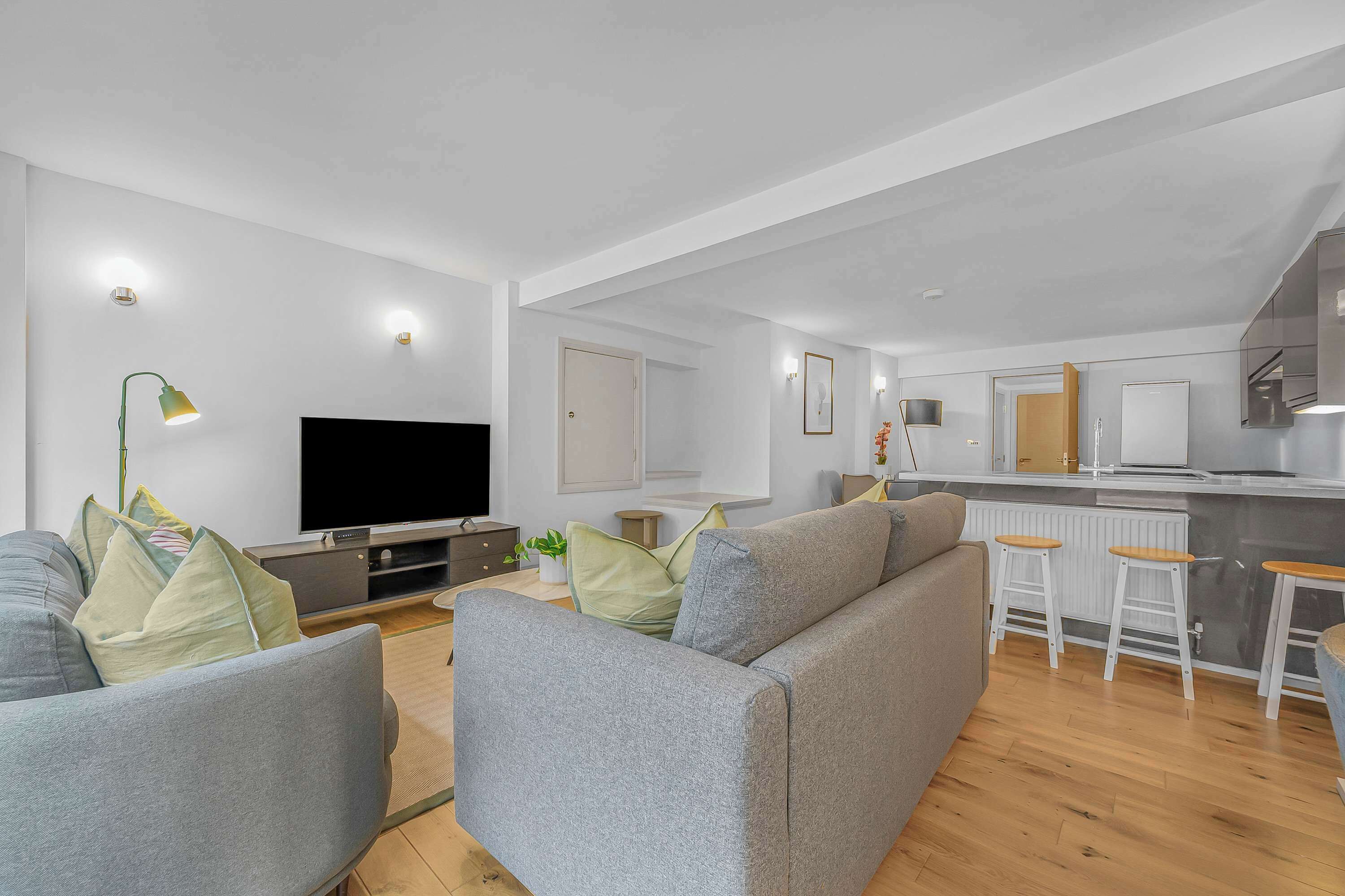 A modern living room with a grey sofa, a flat-screen TV, and a kitchen area with white bar stools.