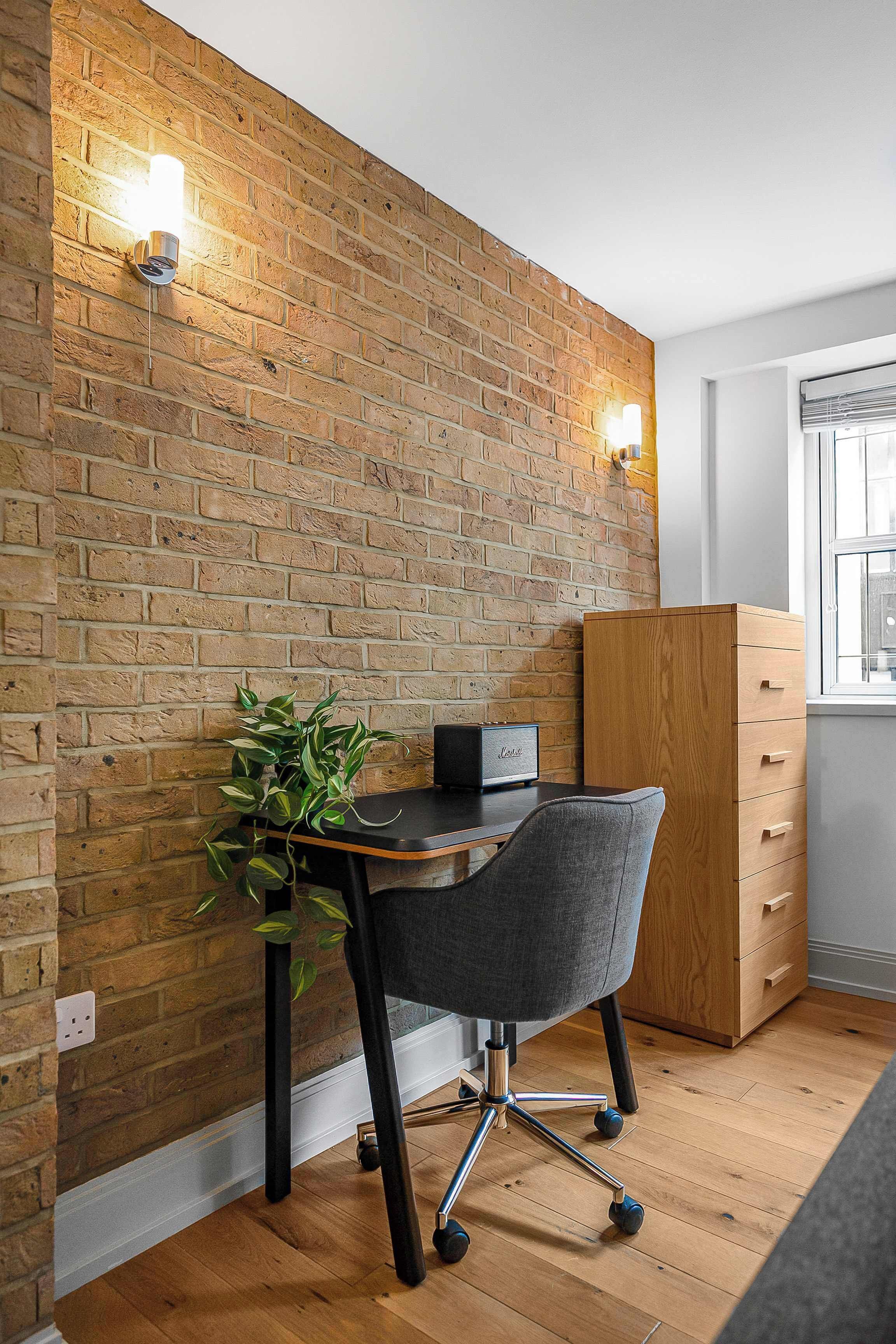A room with a brick wall, a wooden desk, a grey office chair, and a wooden cabinet.