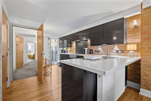 A modern kitchen with dark cabinets, a central island, and a brick accent wall. - Premium 1-bed Apartment in Soho, Near Carnaby St (London)